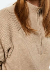 Beige geribbelde gebreide trui met een korte rits aan de voorkant, gedragen door een persoon met lang kastanjebruin haar.