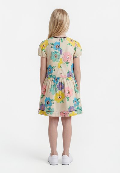 Jeune fille debout, de dos, portant une robe beige avec de grandes impressions florales colorées et des baskets blanches sur fond blanc.