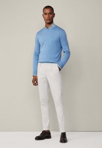 Hellblau gestrickter Pullover, weiße maßgeschneiderte Hose und dunkelbraune Lederschuhe. Der Pullover hat einen Rundhalsausschnitt; die Hose hat eine glatte Oberfläche.