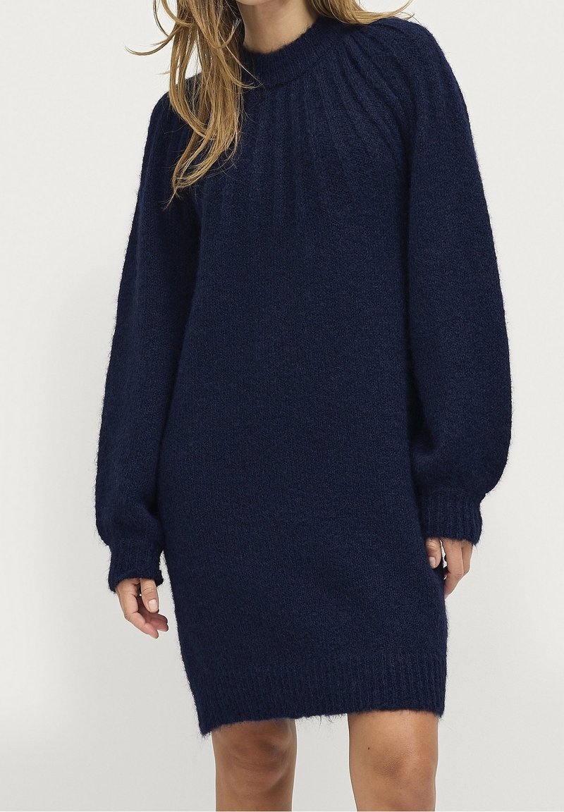 Femme portant une robe pull en laine bleu marine à manches longues avec des poignets, un col et un ourlet côtelés, debout devant un fond uni.