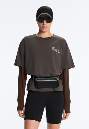 Osoba nosiaca čiernu čiapku, veľké čierne slnečné okuliare, vrstvené hnedé dlhé rukávy a tričko s textom, čierne krátke nohavice a čierne vrecúško na pás.