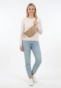 Mujer con suéter rosa claro, jeans azules, zapatillas blancas, sujetando un bolso cruzado beige sobre el pecho, de pie sobre un fondo blanco.