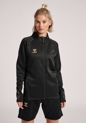 Frau trägt schwarze Sportjacke mit durchgehendem Reißverschluss und passenden Shorts mit goldenen hummel-Logos, steht vor hellem einfarbigem Hintergrund.