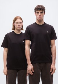 Μαύρα βαμβακερά μπλουζάκια με στρογγυλή λαιμόκοψη, με μικρό λευκό λογότυπο Lacoste στο αριστερό στήθος. Φορεμένα με καφέ παντελόνια από ύφασμα.