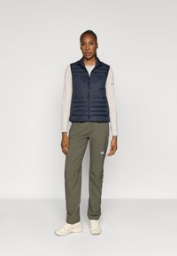 Colete acolchoado leve em azul-marinho com fecho à frente, combinado com calças verde-oliva. Ténis neutros completam o visual. Design minimalista.