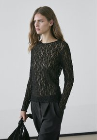 Haut noir en dentelle à manches longues avec un motif texturé à trame ouverte. Il présente un col rond et une silhouette ajustée. Porté avec un pantalon noir.