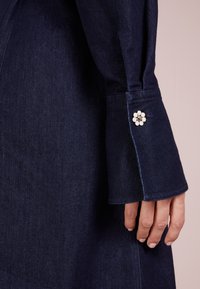 Tumma denim-mekko, jossa pitkät hihat ja napillinen kalvosin, jossa valkoisella ja hopeisilla yksityiskohdilla koristeltu kukkakuvio. Sileä pinta.