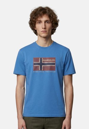 Jeune homme aux cheveux bouclés portant un t-shirt bleu avec un drapeau rectangulaire et le texte "Research Program" sur le devant.