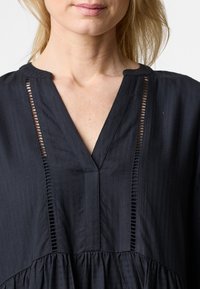 Schwarzes Kleid mit V-Ausschnitt, das vertikale Falten und dekorative Ausschnitte an der Vorderseite aufweist. Weicher Stoff mit strukturiertem Finish.