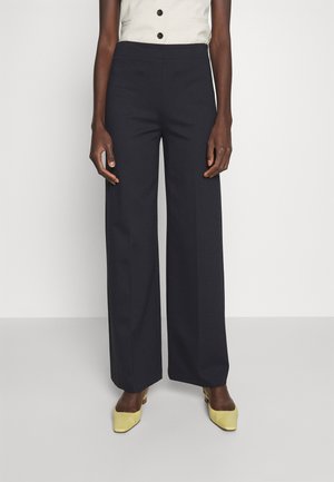 Femme portant un pantalon noir taille haute à jambes larges, un haut blanc sans manches boutonné et des chaussures jaunes à bout pointu, debout devant un fond uni.