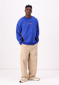 Felpa blu con accenti rossi e pannelli laterali neri, abbinata a pantaloni beige e scarpe da ginnastica color beige. Il modello è in piedi davanti a uno sfondo bianco.