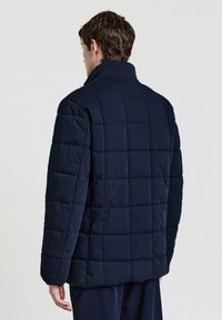 Giacca trapuntata in navy con colletto alto, caratterizzata da motivi a quadrati cuciti e una texture morbida. Adatta per il clima freddo.