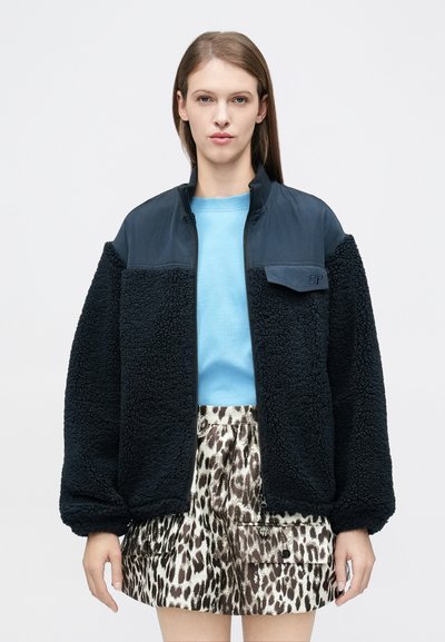 Femme portant une veste en polaire bleu marine sur un haut bleu clair et une jupe à motif léopard marron et blanc, debout devant un fond uni.
