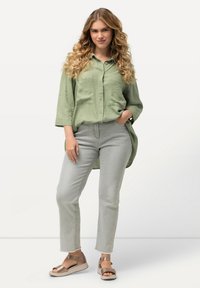 Camicetta verde chiaro con bottoni e due tasche, abbinata a jeans grigi chiari e sandali bronzo con design a fascia.