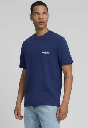 Giovane uomo che indossa una maglietta Replay Inc. blu navy e jeans azzurri, che guarda leggermente verso sinistra contro uno sfondo semplice.