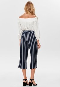 Kvinna klädd i vit off-shoulder-blus, marinblå cropped byxor med vita vertikala ränder och svarta öppna blockklackar, stående med ryggen vänd.