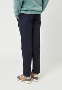 Een persoon staat met de handen in de zakken, gekleed in een marineblauwe broek, een lichtblauwe fleece jas en grijze sneakers met oranje accenten.