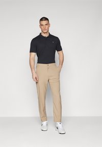 Crna kratka polo majica s ovratnikom, uparen s bež hlačama. Model nosi bijele tenisice; jednostavan dizajn i prilagođeni oblik.