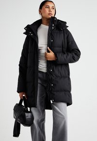 Abrigo de plumas largo y negro con cuello alto, botones a presión y acolchado horizontal; llevado sobre una blusa clara y combinado con vaqueros grises y un bolso negro.