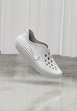 Zapato blanco sin cordones con agujeros de ventilación y logo de Nike, flotando sobre un suelo reflectante con un telón de fondo de cortina luminosa.