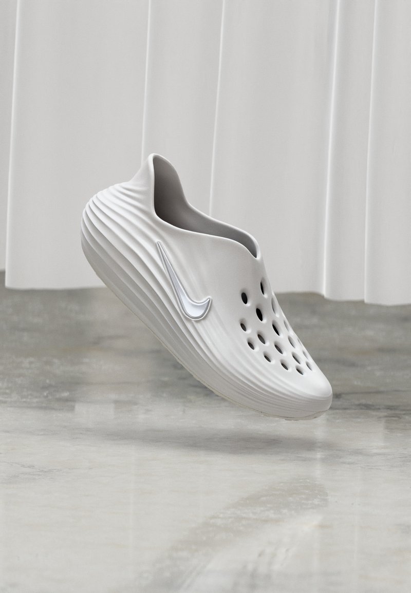 Chaussure blanche sans lacets avec des trous de ventilation et le logo Nike, flottant au-dessus d'un sol réfléchissant avec un rideau lumineux en arrière-plan.