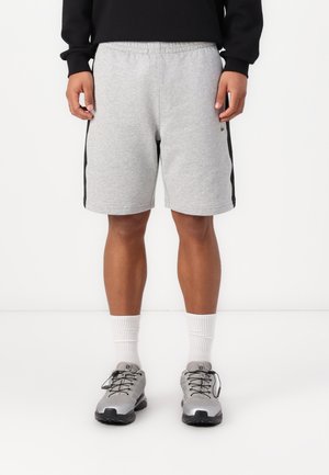 Person trägt hellgraue Sportshorts mit schwarzen Seitenstreifen, weiße Sportsocken, graue Laufschuhe und ein schwarzes Langarmshirt.