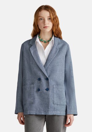 Blazer bleu texturé à double boutonnage avec de grandes poches à rabat, doté de boutons foncés et d'un col châle, associé à une chemise blanche.