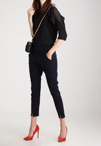 Blouse noire à manches superposées, pantalon marine avec fentes aux chevilles, et escarpins rouges à talons aiguilles. Petit sac bandoulière noir.