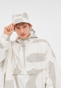 Biela dažďová bunda s veľkými šedými abstraktnými vzormi, zips na prednej strane, elastické manžety a zodpovedajúca vedro čiapka s nášivkou loga.