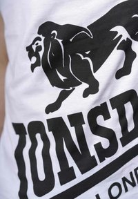 Balta medvilninė marškinė su dideliu juodu liūto grafiku ir ryškiu tekstu. Dizainas turi sklandžias linijas ir ryškius kontrastus.