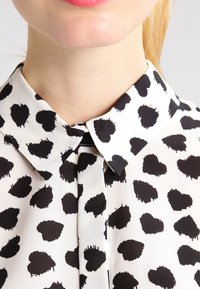 Schwarz-weiße Bluse mit einem Kragendesign und einem Herzmuster. Der Stoff ist glatt, und die Herzen sind unregelmäßig und strukturiert.