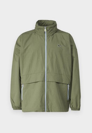 Olive green jacket with a high collar, front zipper, elastic cuffs, and two side pockets; features a small logo on the chest.