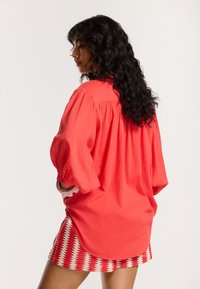 Blusa corallo oversize con maniche a sbuffo, dettaglio arricciato sul retro e vestibilità ampia, abbinata a una gonna a righe rosso e bianco con texture.