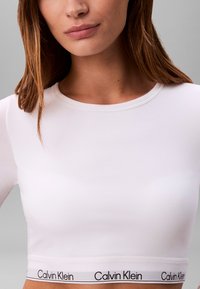 Crop top bianco a maniche lunghe realizzato in materiale liscio, con scollatura arrotondata e un elastico con il marchio Calvin Klein in fondo.