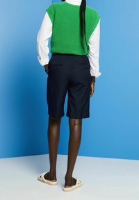 Personne portant un short bleu marine jusqu'aux genoux, une chemise blanche à manches longues, un gilet en maille vert et des sandales beige, debout devant un fond bleu.