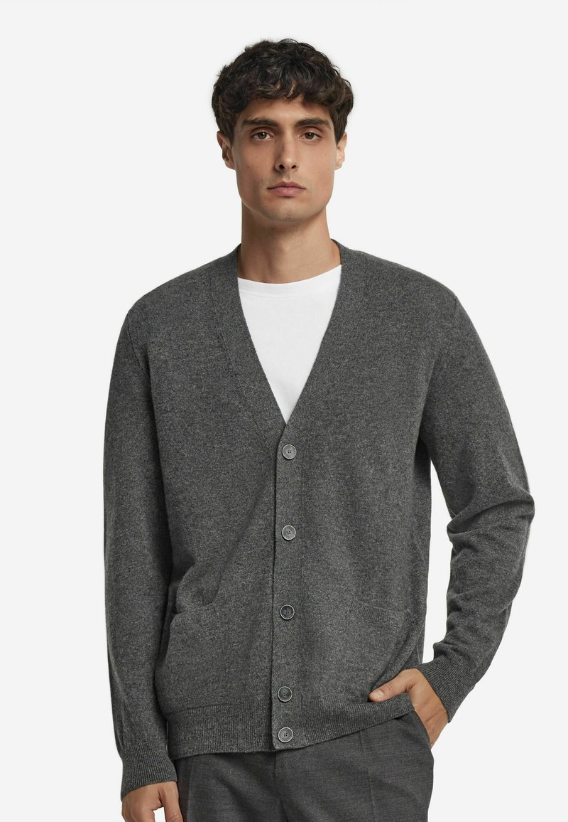 Mężczyzna w szarym kardiganie zapinanym na guziki, noszący białą koszulę, z jedną ręką w kieszeni, stojący na tle jasnego, jednolitego tła.