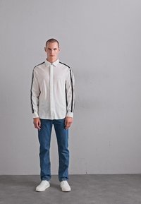 Man som står mot en grå vägg iförd en vit skjorta med svarta ränder på ärmarna, blåjeans och vita sneakers.