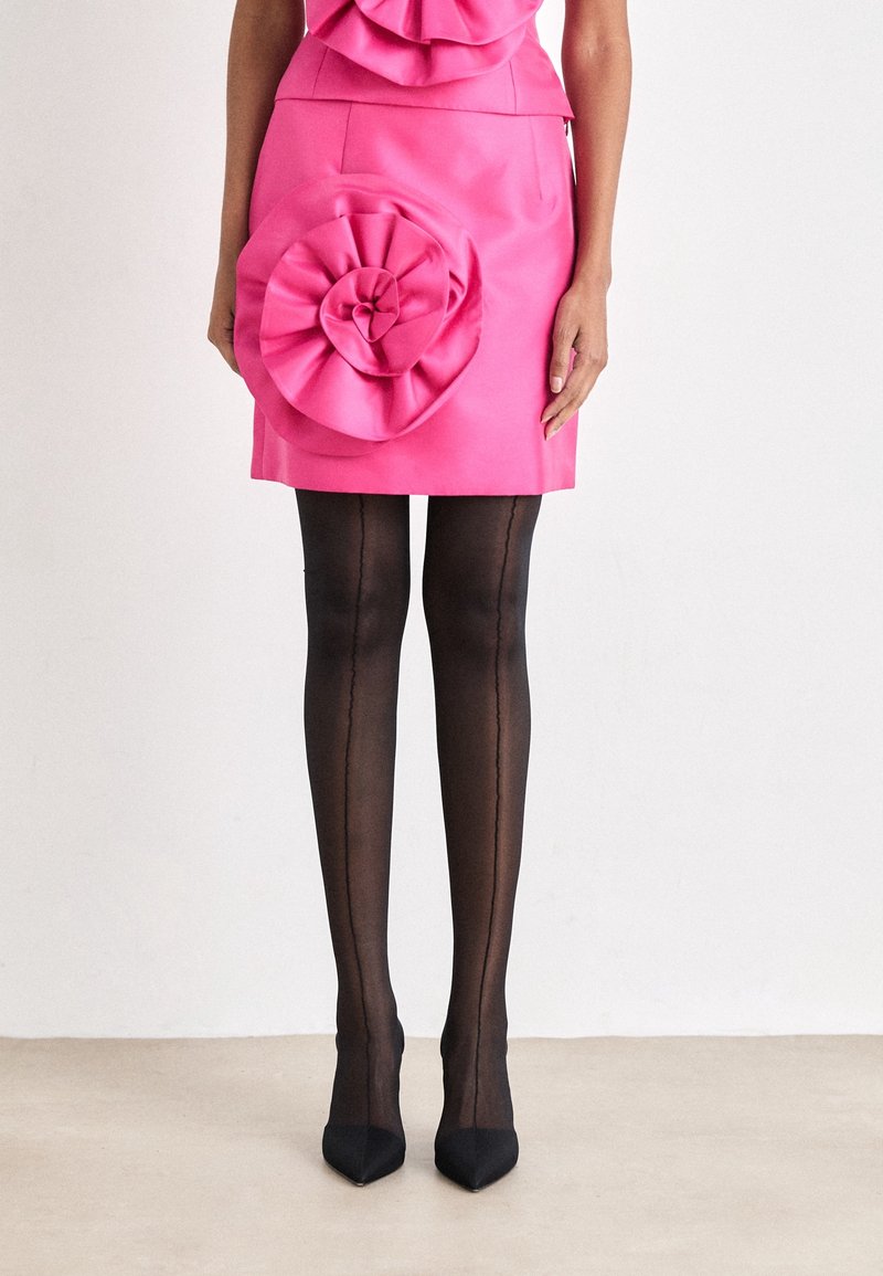 Femme portant une jupe rose vif avec une grande rosette en tissu sur la hanche, associée à des bas noirs à couture et des escarpins noirs pointus.
