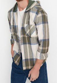 Camicia di flanella a quadri con cappuccio verde, caratterizzata da strisce blu, beige e grigie. Include una tasca sul petto e maniche arrotolate.