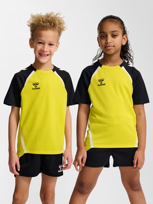 To børn iført matchende gule og sorte sportstrøjer og sorte shorts, der står side om side mod en ensartet baggrund.