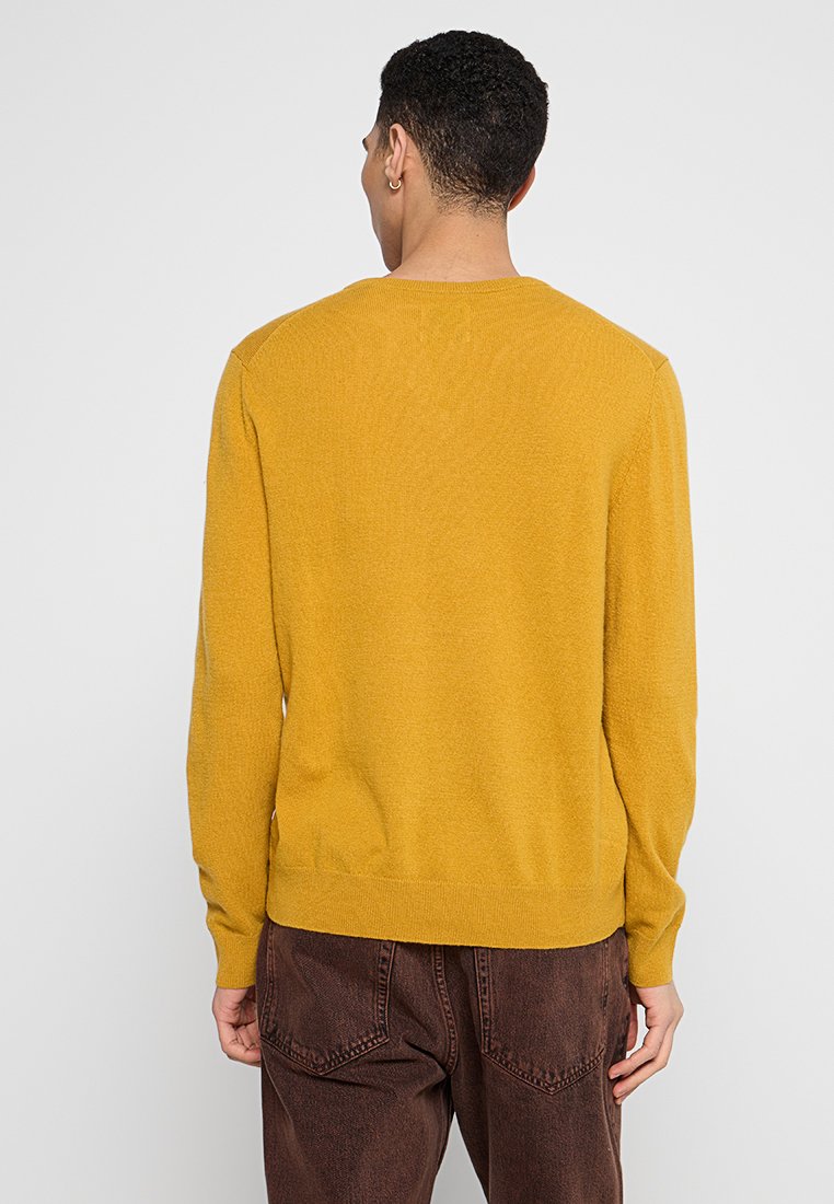 Man med kort lockigt hår som bär en senapsgul långärmad tröja och mörkbruna byxor, vänd bort från kameran mot en enkel bakgrund.