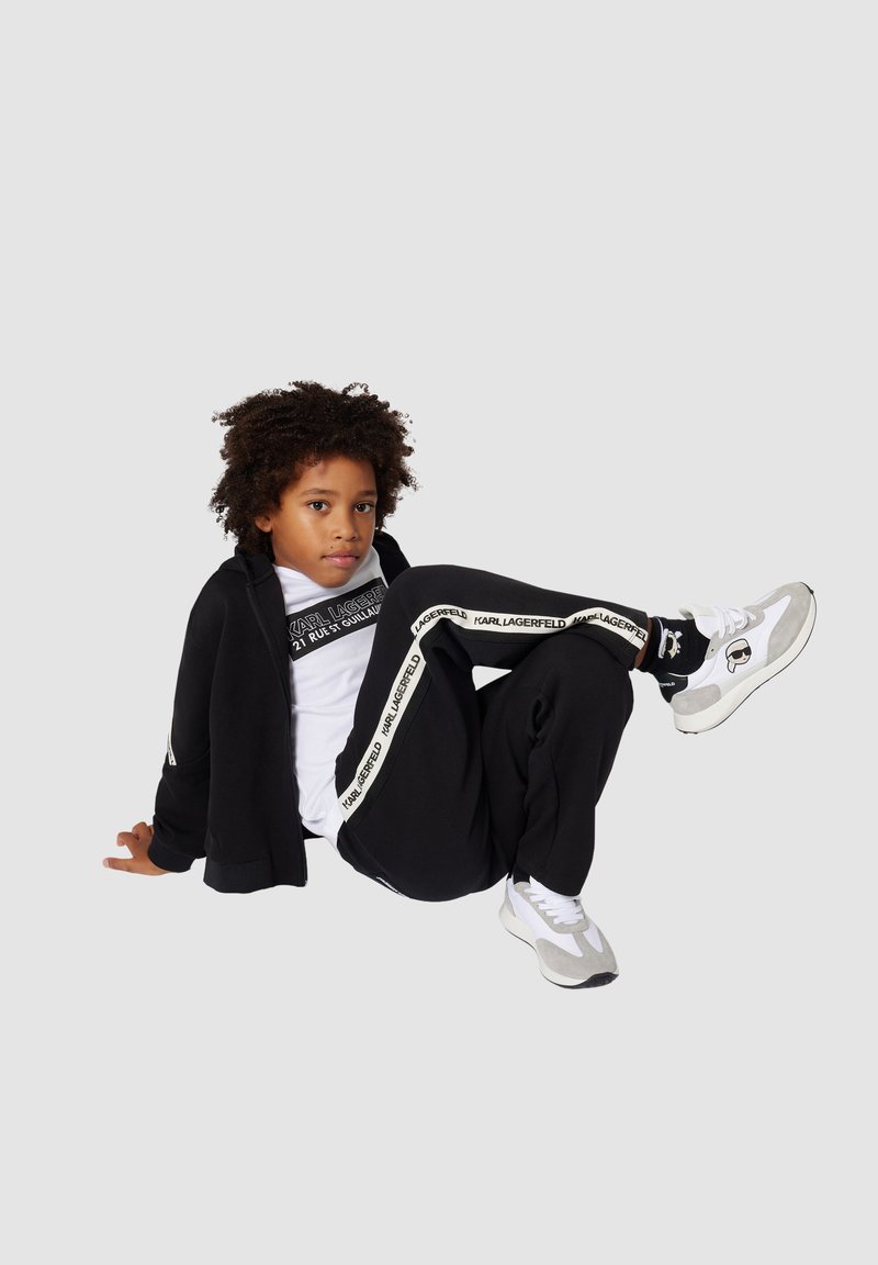 Survêtement noir avec des accents de logo blancs, associé à un t-shirt blanc. Baskets grises et blanches, en pose assise avec des cheveux bouclés.