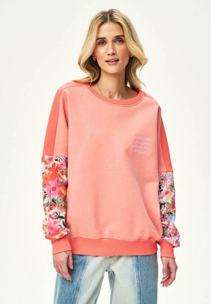 Kobieta ubrana w luźną koralową bluzę z kwiatowym nadrukiem na rękawach i niebieskie dżinsy, zwrócona przodem, o neutralnym wyrazie twarzy.