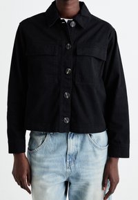 Mustat lyhyt denim-takkia, jossa on kaksi rintataskua, nappikiinnitys ja terävä kaulus. Yllä vaaleansiniset kulutetut farkut.