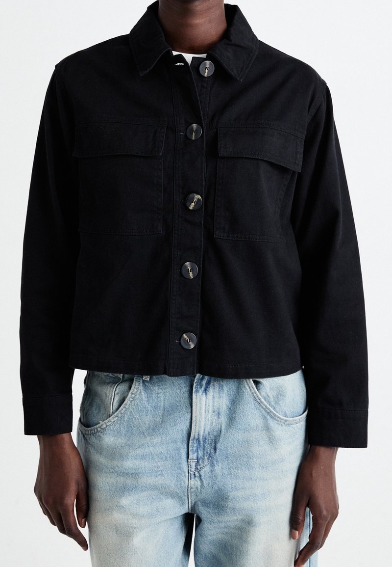 Mustat lyhyt denim-takkia, jossa on kaksi rintataskua, nappikiinnitys ja terävä kaulus. Yllä vaaleansiniset kulutetut farkut.