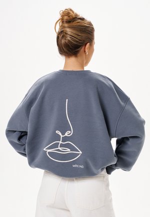 Frau mit Haaren zu einem Dutt gebunden, trägt einen blauen Sweatshirt mit einer weißen abstrakten Linienzeichnung eines Gesichts auf dem Rücken, kombiniert mit hellen Hosen.