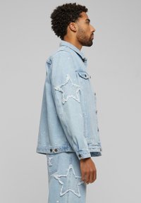 Light blue denim jacket with distressed star patches, button front, and subtle white paint splatters; textured fabric with a relaxed fit.