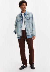 Jachetă din denim de culoare albastru deschis, cu închidere cu nasturi și buzunare frontale, purtată cu pantaloni maro închis și pantofi negri, deasupra unei cămăși albe.
