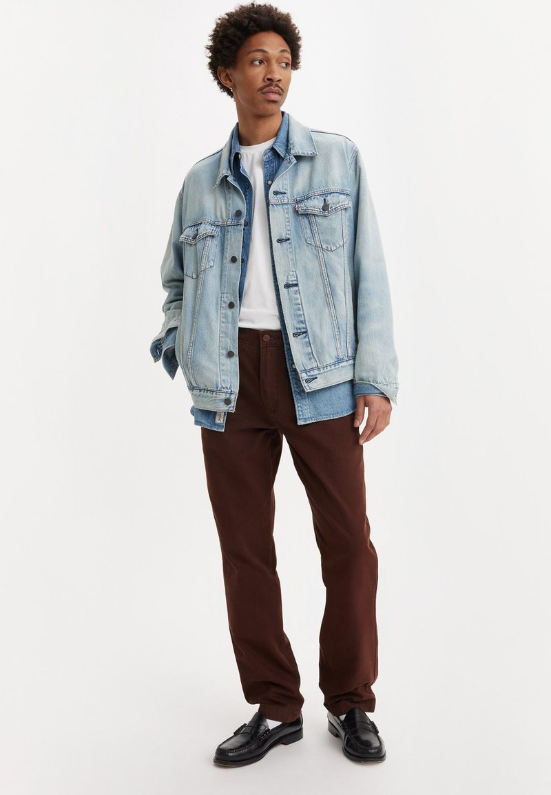 Giacca di jeans azzurra chiara con chiusura a bottoni e tasche frontali, abbinata a pantaloni marroni scuri e scarpe nere, indossata sopra una camicia bianca.