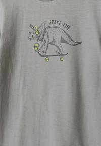 Grå T-shirt med en illustreret triceratops på et skateboard, der har høretelefoner på og holder en smartphone. Teksten siger "SKATE LIFE."